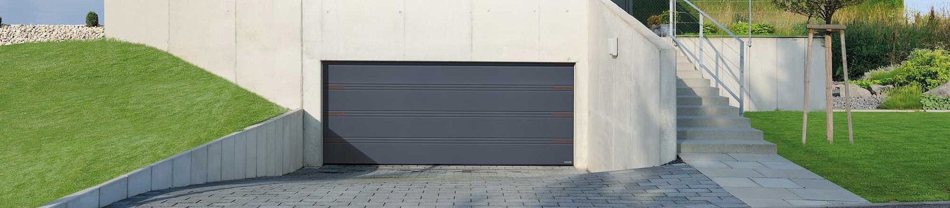 Einfahrt mit geschlossenem, dunkelgrauem Garagentor neben einer Treppe mit Edelstahlgeländer und gepflegtem Rasen
